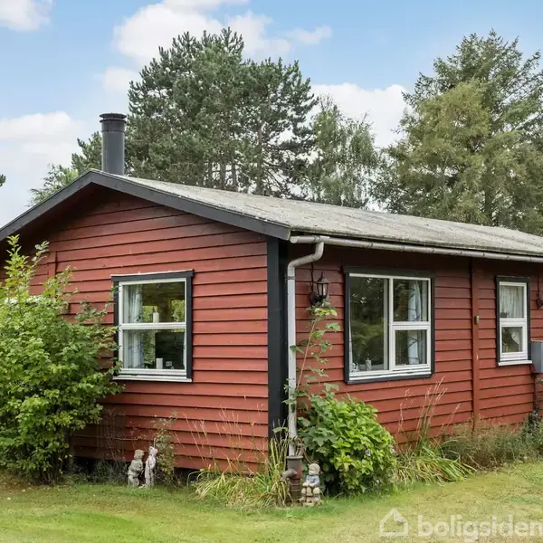 Et rødt træhus står stille omgivet af grønne buske og græs. Små vinduer og en skorsten vises. Baggrunden består af tætte træer og en blå himmel med få skyer.
