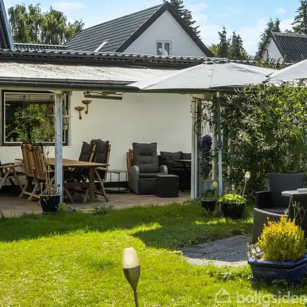 Havemøbler står under en overdækket terrasse med træbord og stole. En stor parasol giver skygge, omgivet af grøn græsplæne og buske. Bagved ses et hvidt hus med sorte tagplader.