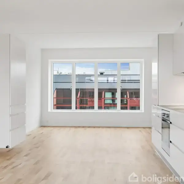 Et lyst køkken-alrum med hvide skabe og træparketgulv. Et stort vindue viser udsigt til nabobygninger og himmel. Rummeligt og minimalistisk design.