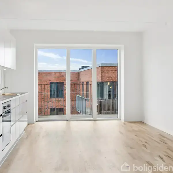 Et lyst køkkenalrum med hvide elementer og trælaminatgulv. Store vinduer fører ud til en altan med udsigt til en rød murstensbygning. Klar, blå himmel i baggrunden.