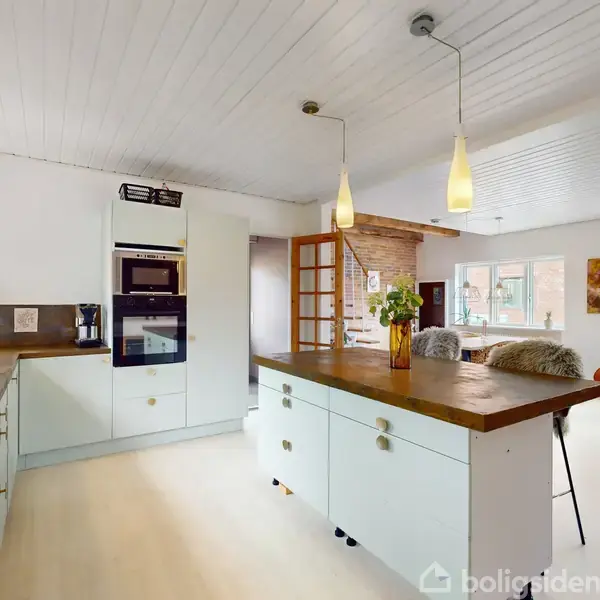 Køkken med hvidt interiør, indbygget ovn og brune træborde. Rummelig stue i baggrunden med sofa, guitar i hjørnet og billeder på væggen. Lyse lofts- og gulvlamper.
