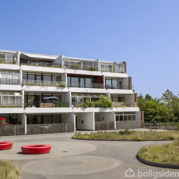 Et moderne lejlighedskompleks med altaner og planter på flere etager. Bygningen står i en solrig, grøn omgivelse med en klar blå himmel som baggrund.