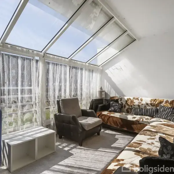 Sofagruppe står i en rummelig stue med store vinduer og lette gardiner, der slipper lys ind. Sollys belyser rummet, hvor også en lænestol og kuffert er placeret.