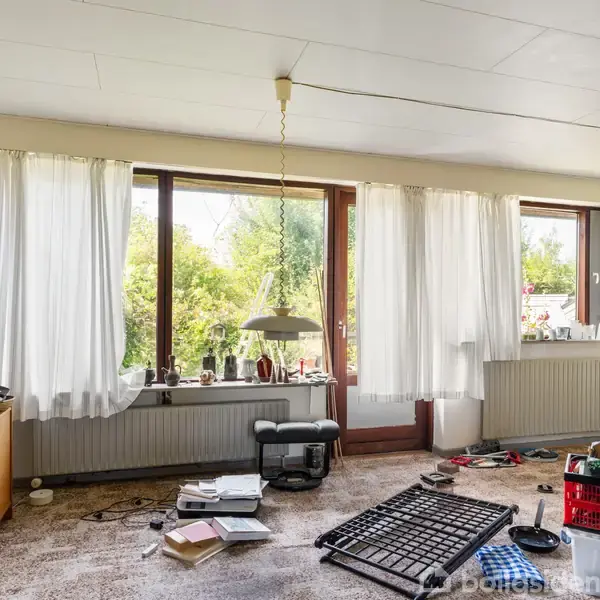 Et rodet stueområde med bøger, en åben reol og diverse genstande spredt på gulvet. Store vinduer med hvide gardiner lukker lys ind, og der ses en grøn have udenfor.