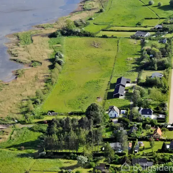 En række huse ligger tæt ved en vej i et frodigt grøn område langs en søbred. Omkringliggende marker og træer giver en landlig atmosfære.