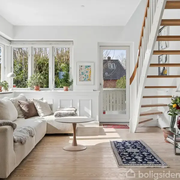 En lys stue med en beige sofa fyldt med puder, en rund hvid bord, trappe til første sal, og vinduer med planter. En brændeovn står ved et lille tæppe. Ingen tekst.