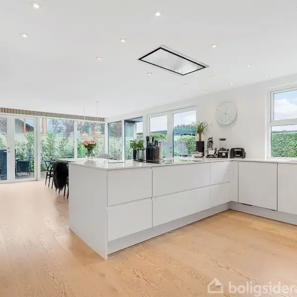Et moderne køkken med hvide skabe fyldt med køkkenudstyr. Store vinduer og glasdøre giver udsigt til en grøn have. Lyse trægulve og minimalistisk design præger rummet.