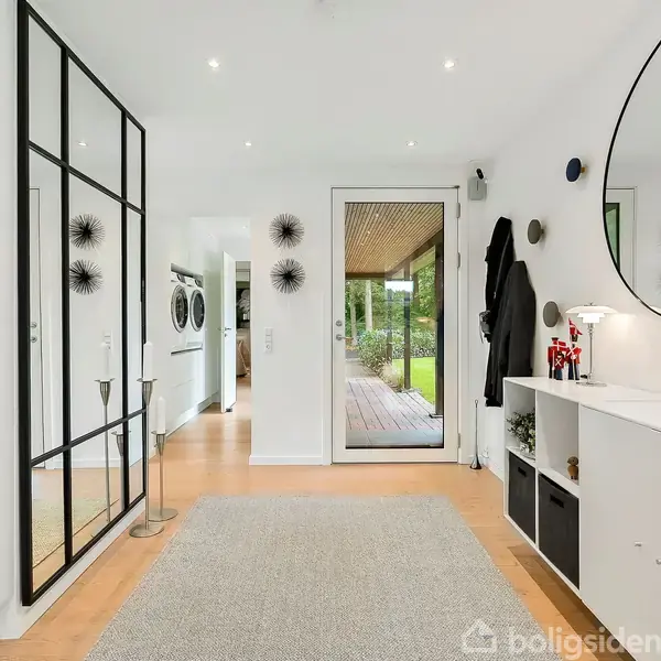 Gang med spejl i sort ramme, skærer sig elegant ind i rummet. Hylde med dekorationer står under cirkulært spejl. Dør fører ud til overdækket terrasse med udsigt til haven.