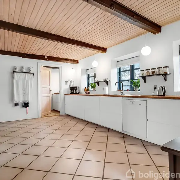 Et moderne køkken med hvide skabe og lyse fliser. Bordplader i træ, vask med armatur, og vinduer med udsigt til træer. Loftet har træpaneler og synlige bjælker.