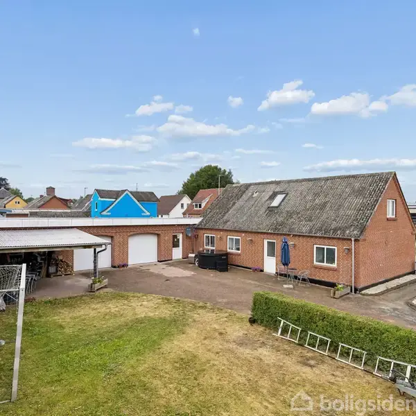 En rød murstenshus med skråt tag står i en gård. Husets omgivelser inkluderer en garage med carport og græsplæne. Blå himmel med få skyer i baggrunden.