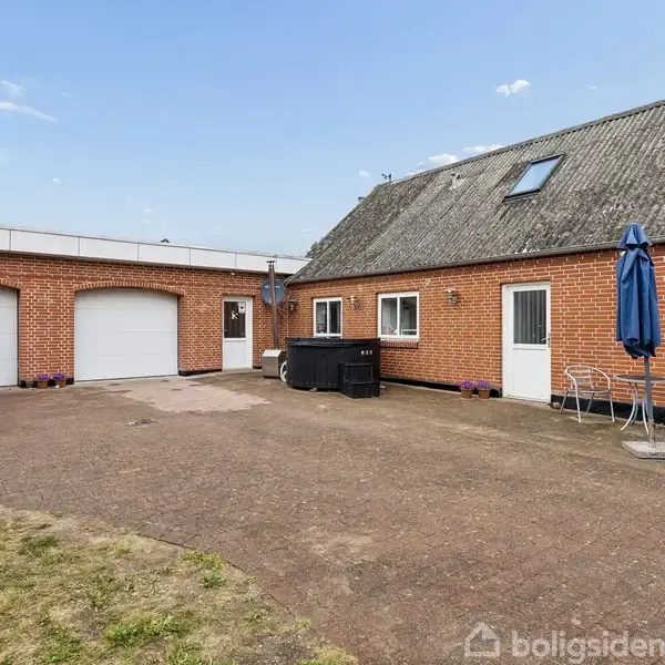 Murstenshus med to garager og skråt tag. Foran huset står et cafébord med stole og en blå parasol. Området er belagt med fliser og omgivet af græsplæne.