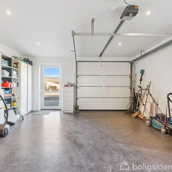 En garage med en åben, udtrækkelig port. I venstre side er der reoler og skabe med værktøj. Højre side har stablede dæk og haveredskaber. Gennem en dør ses dagslys udefra.