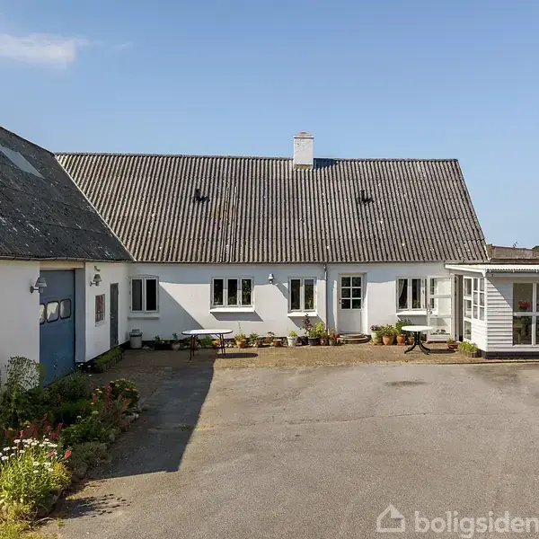 Hus med hvid facade og gråt tag står i sollys. Der er en indkørsel foran og blomsterbed langs bygningens venstre side. En lille udestue er til højre.