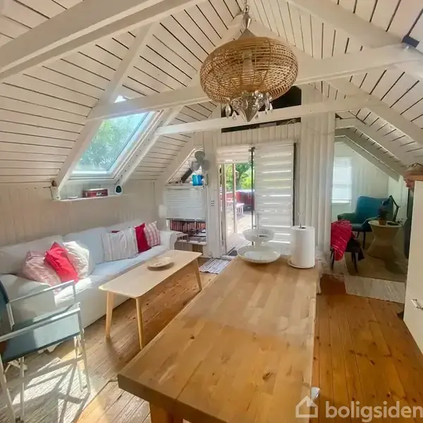 Stue med hvid sofa pyntet med farvede puder, stående under skråvindue, træbord i midten, og udgang til terrasse gennem glasdør. Indretningen er lys og hyggelig.