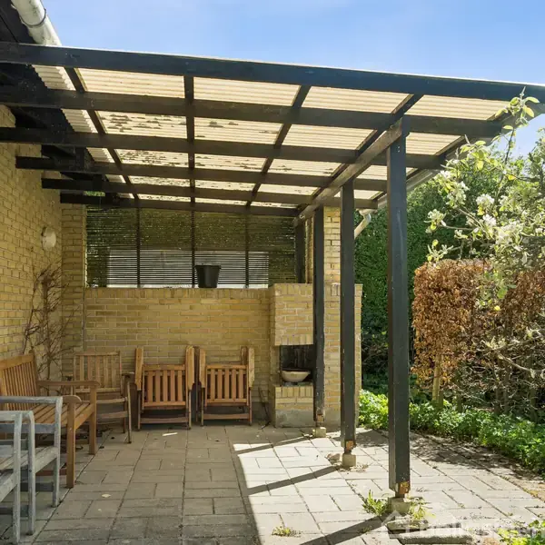 Terrasse med trægulv er møbleret med træstole og et lille bord. Tagkonstruktion af gennemsigtigt materiale giver skygge. Omkranset af grønne planter og buske i et solrigt udendørs miljø.