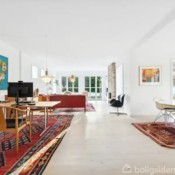 Et lyst, moderne rum med et skrivebord og computer til venstre, en spiseplads til højre, røde tæpper under møblerne, og store vinduer, der giver naturligt lys.