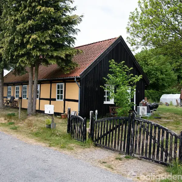 Hus med sort bindingsværk og rødt tegltag står ved en vej. Træer og buske omgiver huset, mens en åben træport fører til en have med bil i baggrunden.