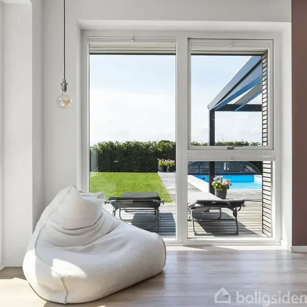 En stor, hvid sækkestol står i et lyst rum foran en glasdør, som åbner ud til en terrasse med pool, liggestole og en velplejet græsplæne under en klar himmel.
