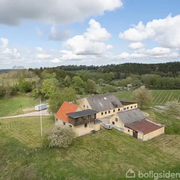 En gårdbygning med rødt tag og gul facade står på en stor græsplæne omgivet af marker og skov. Himlen er let skyet, og der er få træer omkring.