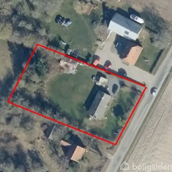 En aflang grund indhegnet med en rød streg. På grunden er der et hovedhus omgivet af græs, nogle mindre bygninger og parkerede biler. Vejen løber til højre.