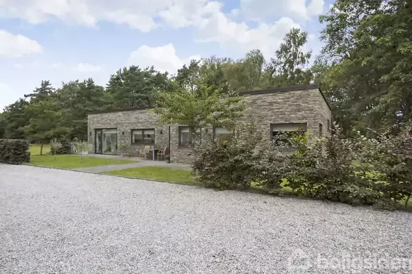 En moderne bungalow i mursten ligger stille i en grøn have, omgivet af tætte buske og træer, med en grussti foran.