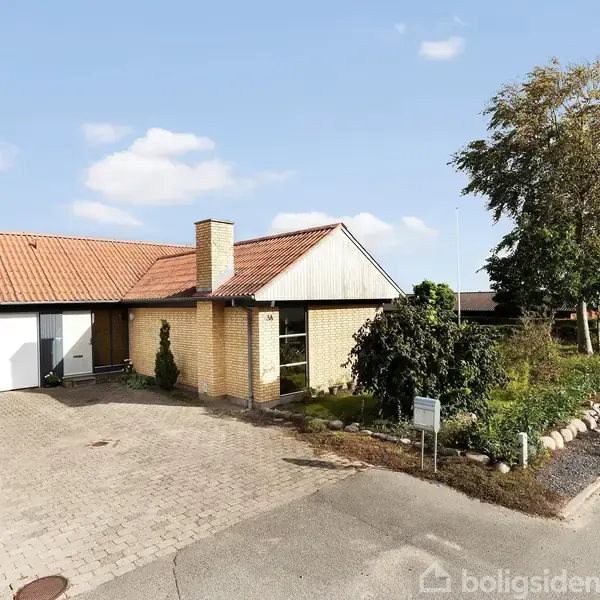 En etplans hus med rødt tegltag og garage står stille i en boligkvarter. Forhaven har buske og træer omkranset af sten. Skilt står ved indkørslen.