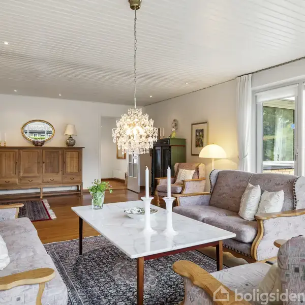 En sofagruppe med puder står omkring et rektangulært, hvidt sofabord. Rummet har en lysekrone, træskab og tæppe. Ved siden af er der store vinduer med udsigt til en terrasse.