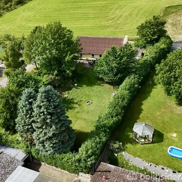 Grøn græsplæne omgivet af høje træer og buske. Midt på græsset står et lille lysthus og et opstillet soppebassin. Omkring ses en gård og marker.