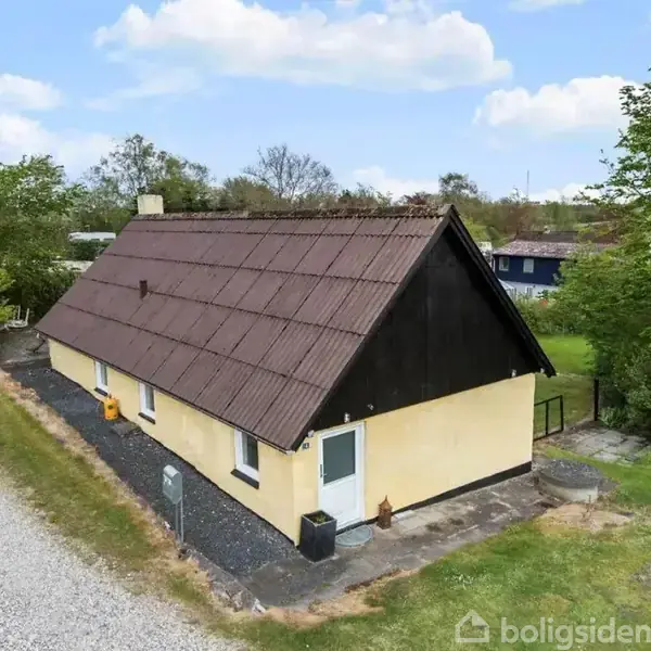 Hus med gult ydre og brunt tag står stille ved en grusvej. Omkring huset er der græs og buske under en blå himmel med skyer.