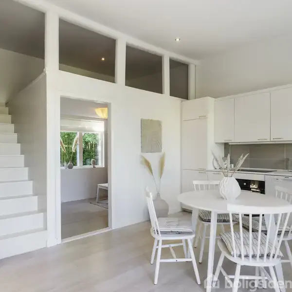 Et lyst køkken-alrum med hvidt spisebord og stole, ved siden af et moderne køkken. En hvid trappe leder til en overetage. Rummet er minimalistisk indrettet med neutrale farver.