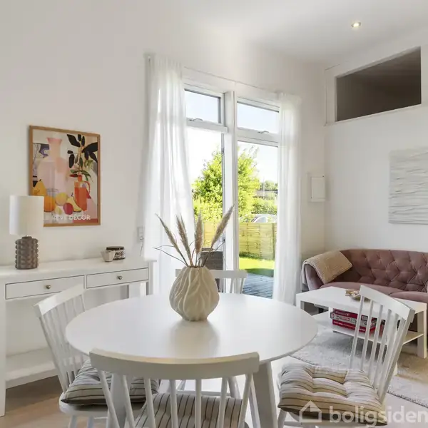 Rundt spisebord med vase og stole står i stue, nær sofa og kunst. Sollys strømmer ind fra stor havedør, oplyser lys væg farver og skaber hyggelig atmosfære.