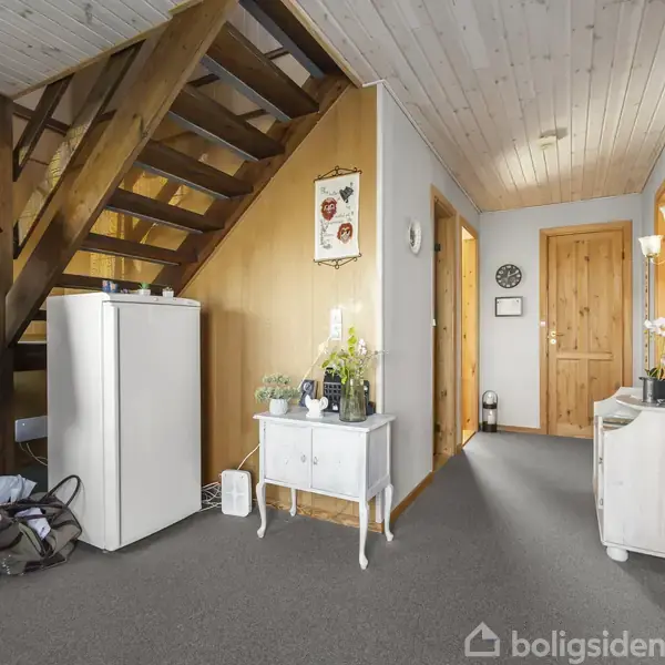 En entré med en trappe, jakker hængende på en knagerække, og et lille hvidt skab. Et tæppe dækker gulvet, og flere døre fører til andre rum.