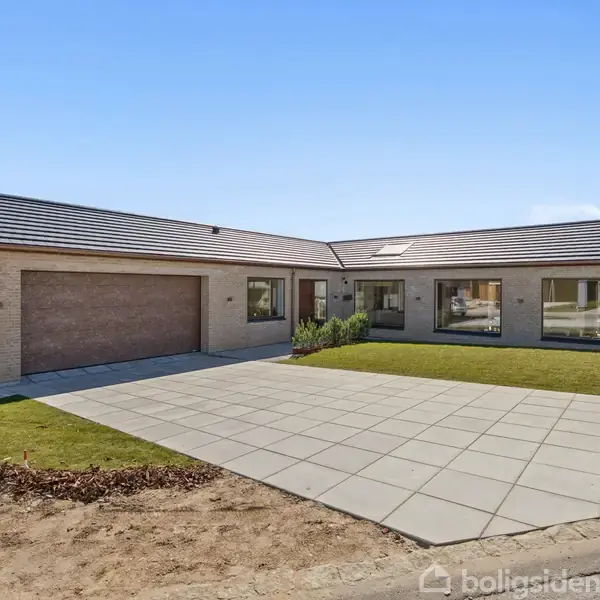 En moderne, vinkelformet etplansvilla med sadeltag, store vinduer og muret facade. Huset står på en flisebelagt indkørsel, omgivet af en nyligt anlagt græsplæne under en klar blå himmel.