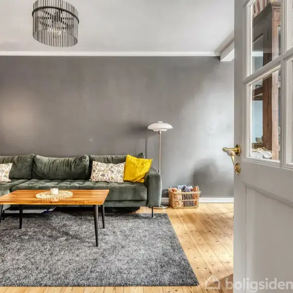 Grøn hjørnesofa med gule puder står i en stue med grå vægge. Sofaen er placeret på et gråt tæppe foran et træbord, ved siden af en hvid standerlampe.