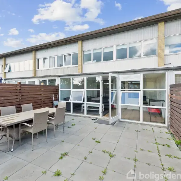 En rækkehus baggård med en fliseterrasse, hvor et bord og seks stole står placeret. Omgivet af trægærder, fører terrassen til en glasindgang med udsigt til en bolig.
