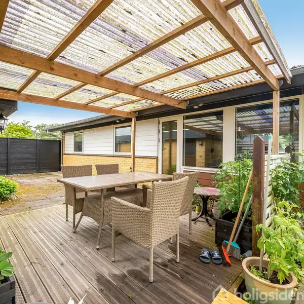 Terrasse med bord og stole under en gennemsigtig overdækning. Omkring stå planter i potter. Bagerst ses et gul murstenshus med store vinduer, og i baggrunden er en sort låge.