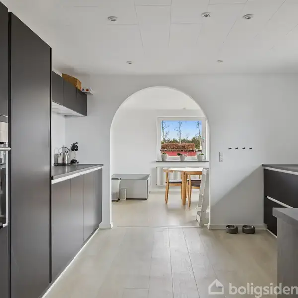 Køkken med sorte skabe og indbygget ovn, forbundet til en spisestue gennem en hvid bue. Store vinduer i baggrunden giver udsigt til træer og blå himmel.