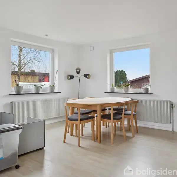 Et rundt spisebord med fire stole står i et lyst hjørne af et værelse med to vinduer. Små planter dekorerer vindueskarmene, og en sort standerlampe står ved siden af bordet.