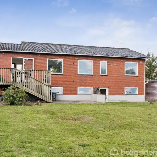 Rødstenshus står stille i en velholdt have med græsplæne. Huset har en trappe til en altan og flere vinduer. Baggrunden er en blå himmel med få skyer.