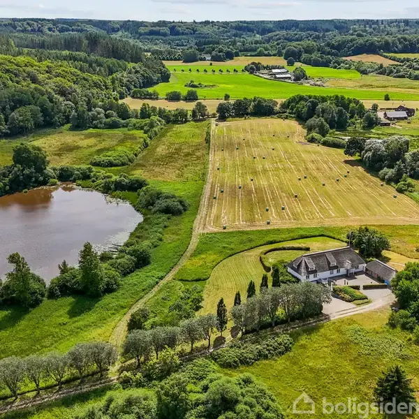 Hus ligger stille ved siden af en mark med rundballer og en sø. Omgivet af frodige grønne marker og skove, der strækker sig i horisonten under en blå himmel.