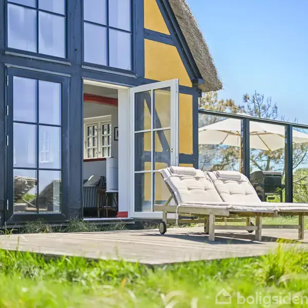 Solstol står på en terrasse foran et bindingsværkshus med gule vægge. Døren til huset står åben, og et glasset vinterhaveområde kan ses i baggrunden.