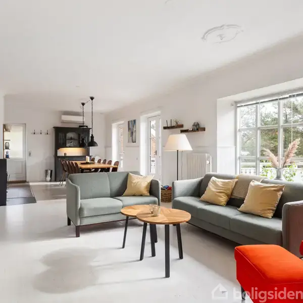 En lys stue med moderne møbler, inklusive en grøn sofa og rød stol. Store vinduer lukker naturligt lys ind. I baggrunden ses en spiseplads med bord og stole.
