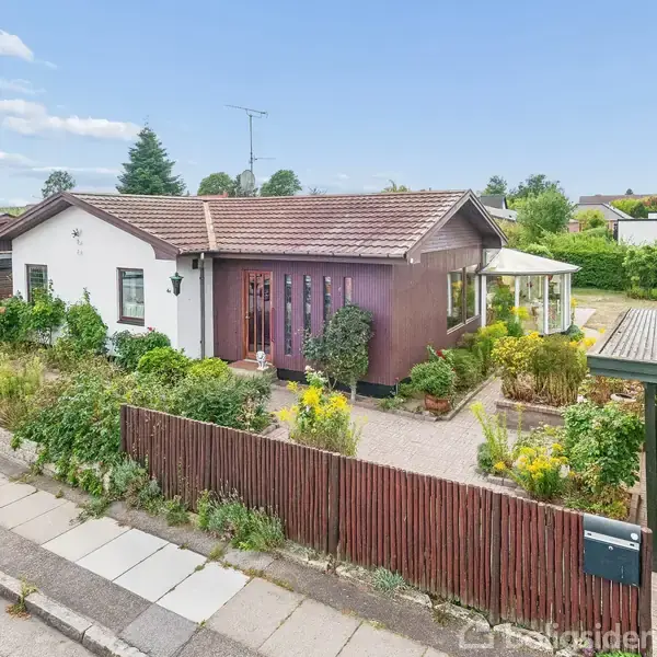 Hus med rødt tag står stille omgivet af have med blomster, beliggende i et villakvarter. Grussti fører til hoveddør, indhegnet af lavt, brunt træhegn.