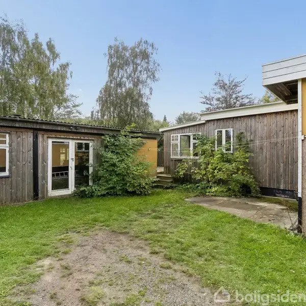 En træbygning står stille i en grøn have med buske og træer i baggrunden. Bygningen har flere vinduer og en glasdør. Græsplænen er delvist bar.