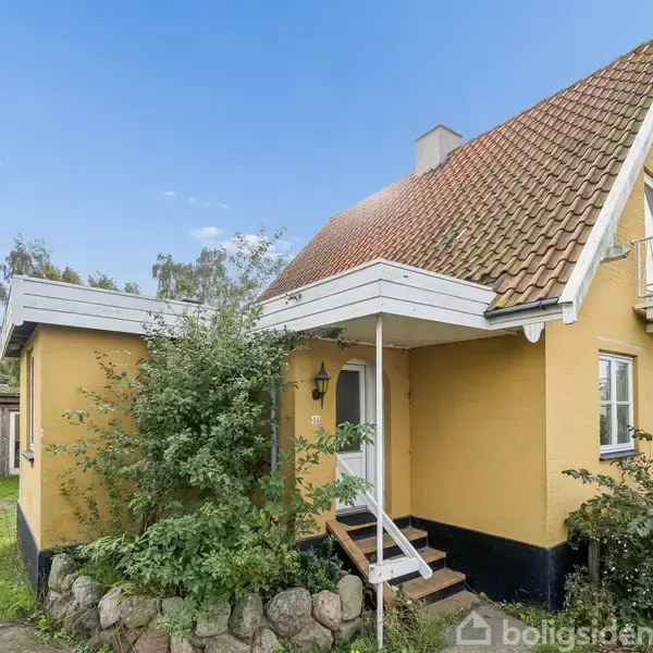 Et gulmalet hus med rødligt tegltag, lille balkon og en trappe ved hoveddøren. Det står i en have med grønne buske, en sti og en mindre bygning i baggrunden.