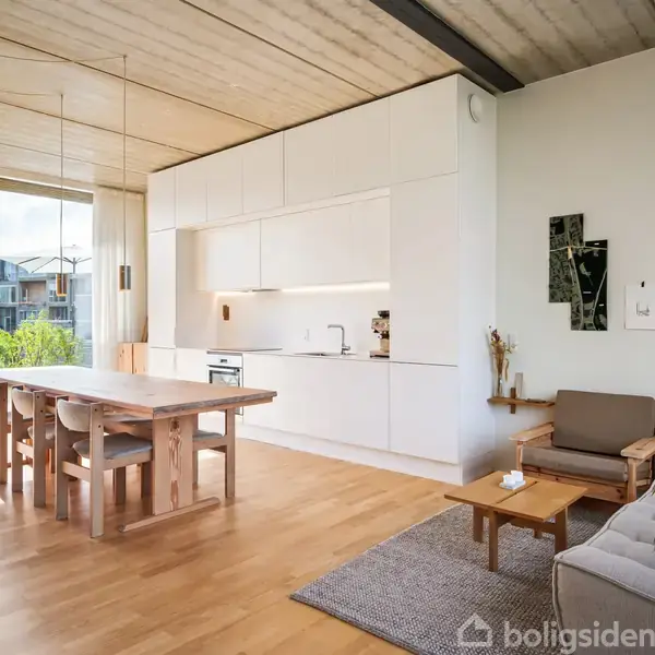 En lys, moderne stue med trægulv og indbygget køkken. Et langt spisebord med stole står ved glasvægge, som åbner ud til en balkon og udsigt. En sofa og lænestol står på et tæppe, ved siden af en unik lampe og vægkunst.