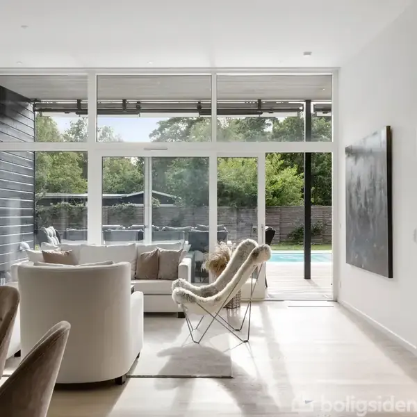 Lys stue med komfortable stole og bord, der vender mod store vinduer med udsigt til en have med pool. Moderne indretning og maleri på væggen.