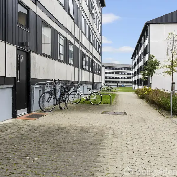 Lejlighedskompleks med to etagers bygninger har cykler parkeret udenfor. En sti løber mellem bygningerne, omgivet af græsplæner og buske. Solskinsvejr og blå himmel i baggrunden.