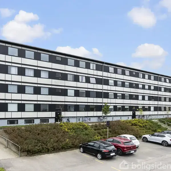 En fler-etagers bygning står stille foran en parkeringsplads, hvor flere biler holder parkeret. I baggrunden ses en grøn beplantning og en blå skyfyldt himmel.