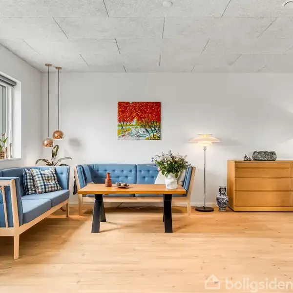 Blåt sofaarrangement står i en stue med lyse trægulve og hvidmalede vægge. En farverig maleri hænger over sofaen. Blomster og lamper pynter bordet og en skænk.